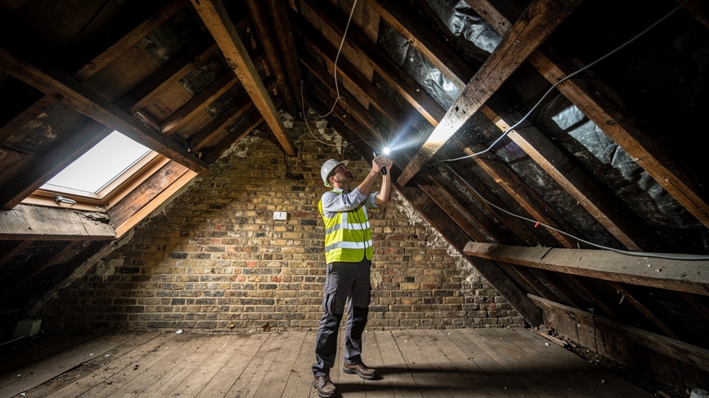 Chartered surveyor with torch inspecting roof space and timber beams in a Victorian London house loft